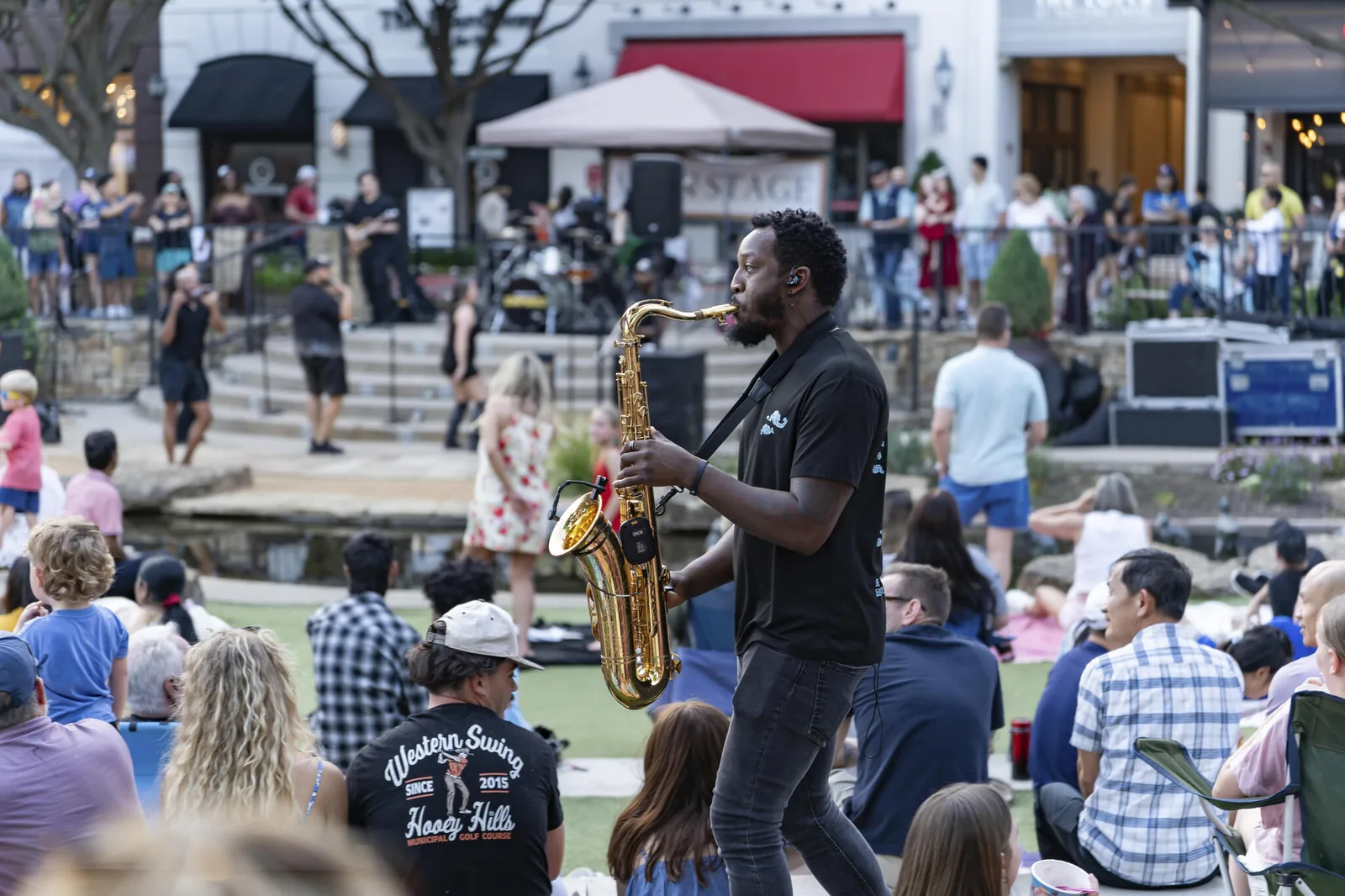 A saxophonist plays among the audience with the festival stage in the background