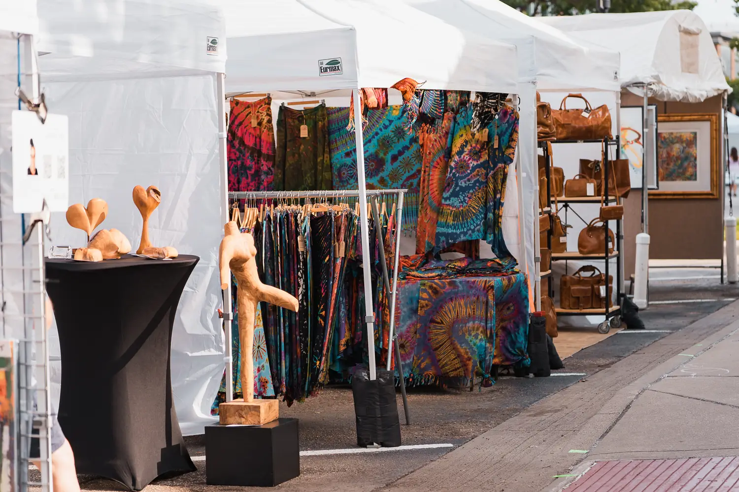 A row of white festival tents with tie-dyed textiles, wood sculpture, and leather bags