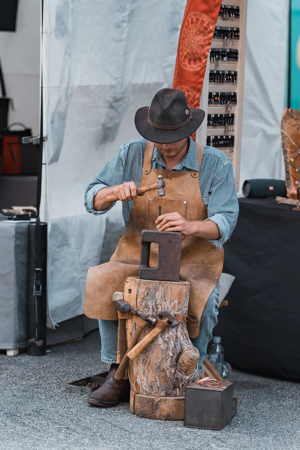 A metalsmith in a leather apron hammers jewelry at an anvil in his festival booth